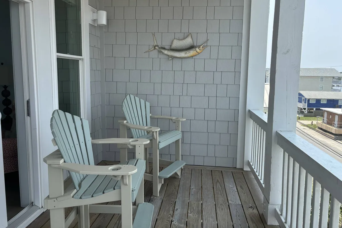 Two light blue Adirondack chairs are positioned against an exterior wall decorated with a fish motif, inviting guests to relax and socialize while enjoying the coastal ambiance. The setup is casual and evokes a laid-back beach vibe.