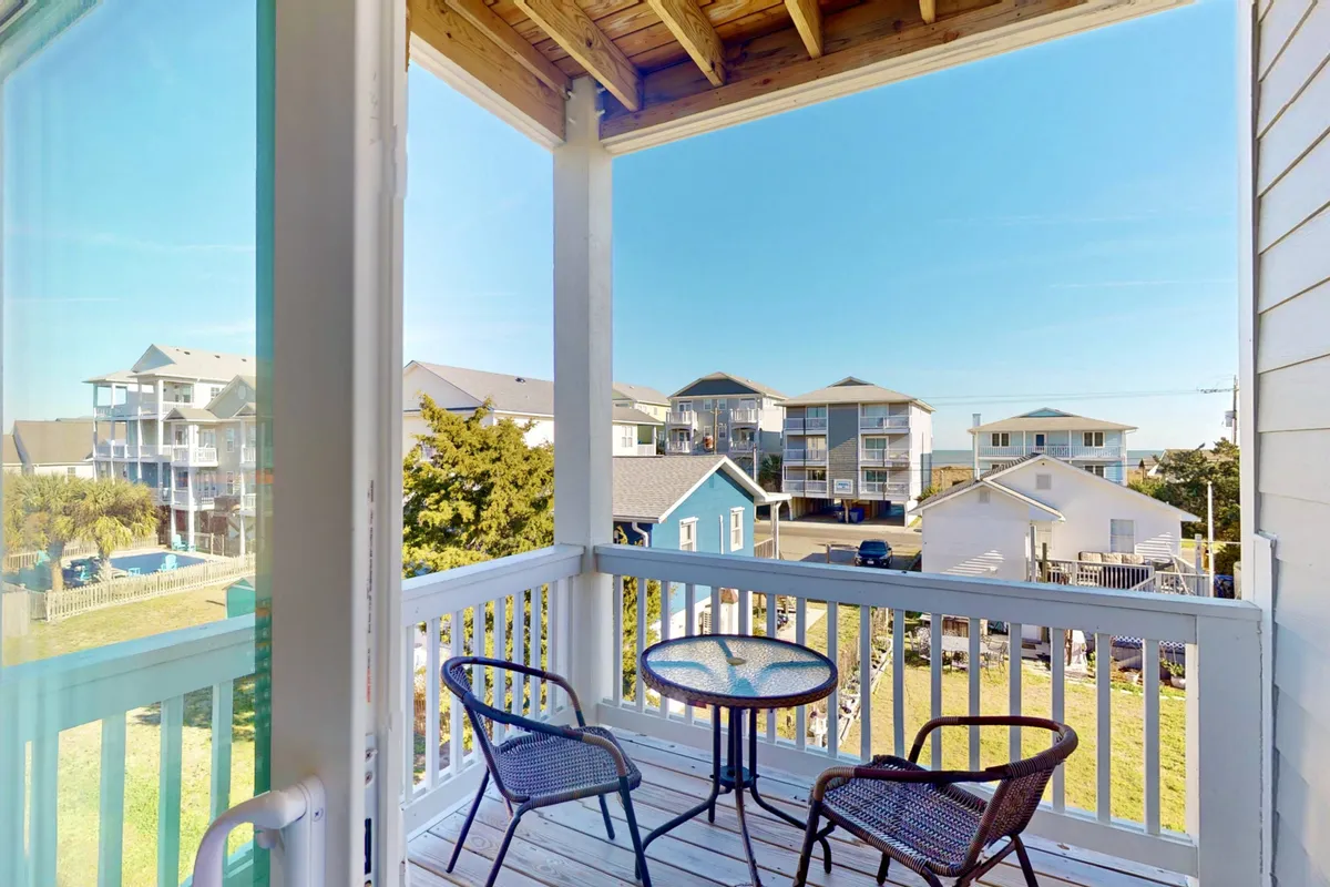 An inviting exterior balcony scene with a table and chairs overlooking the surrounding neighborhood and distant water views. This space is ideal for morning coffee, afternoon relaxation, or taking in the sunset after a day of beachside fun.