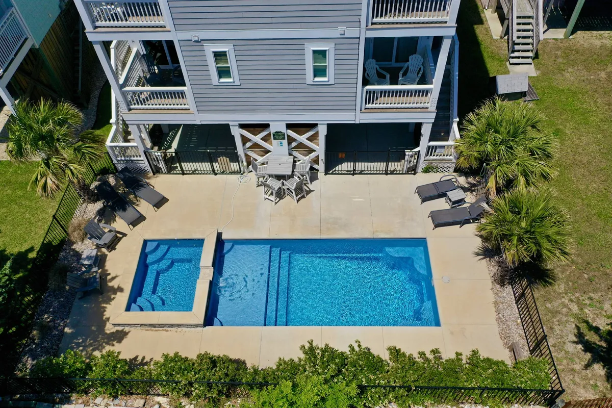 The image showcases the exterior of the Blue Oasis townhome at Carolina Beach, highlighting a sparkling private swimming pool surrounded by inviting outdoor seating and multiple balconies. The scene is set in relaxed coastal light, evoking a sense of leisure and togetherness, with plenty of space for sunbathing, swimming, or gathering poolside with family and friends.