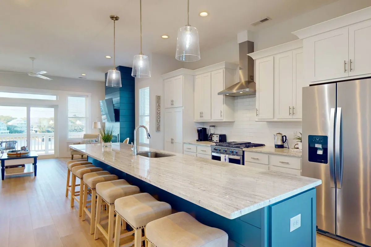 This image features a large kitchen island at the center, surrounded by stainless steel appliances, a bright blue accent wall, and hanging pendant lights. There is ample counter space, stylish bar stools for casual dining, and the space is flooded with natural light, making it ideal for group cooking and entertaining.
