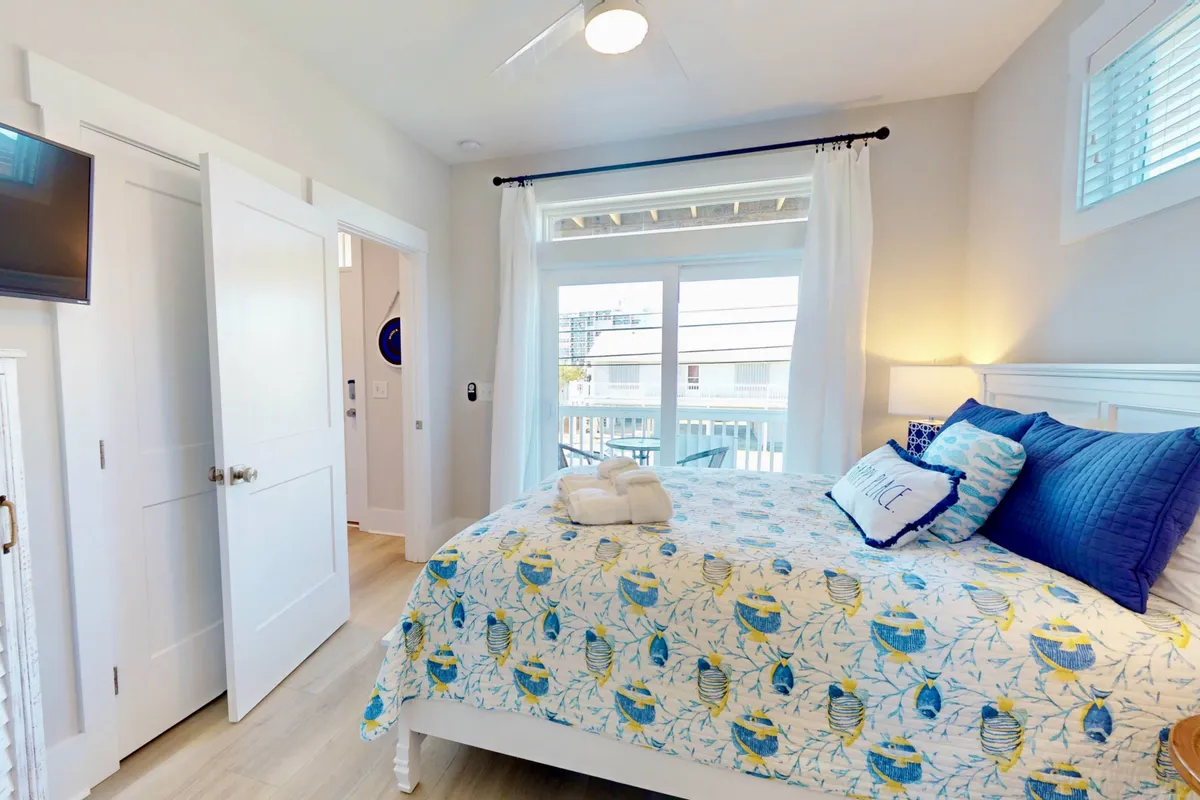 A sun-filled bedroom with a bed featuring a patterned quilt, decorative pillows, and large windows streaming in natural light. The tranquil, cheerful setting feels welcoming and is furnished for relaxation and comfort.