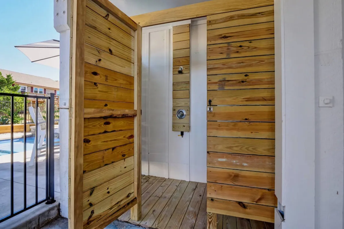 A stylish, wood-accented outdoor shower area is depicted here, with wooden doors, modern shower fixtures, and natural lighting. The scene feels private and rejuvenating, ideal for rinsing off after a day at the beach or pool.