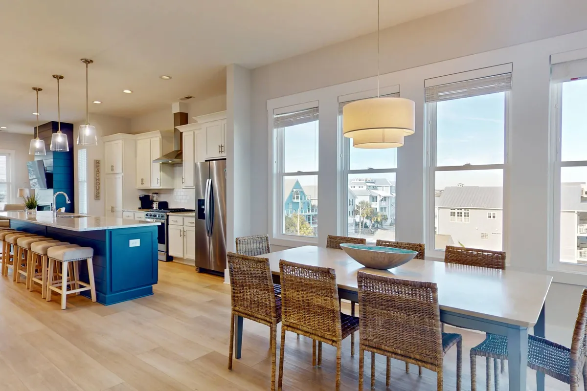 This image showcases an elegant open-plan space with a kitchen island and a large dining table by the window. Sunlight streams through the windows, creating a warm, welcoming ambiance ideal for family meals, celebrations, and socializing. The setup allows for easy flow between cooking and dining areas.