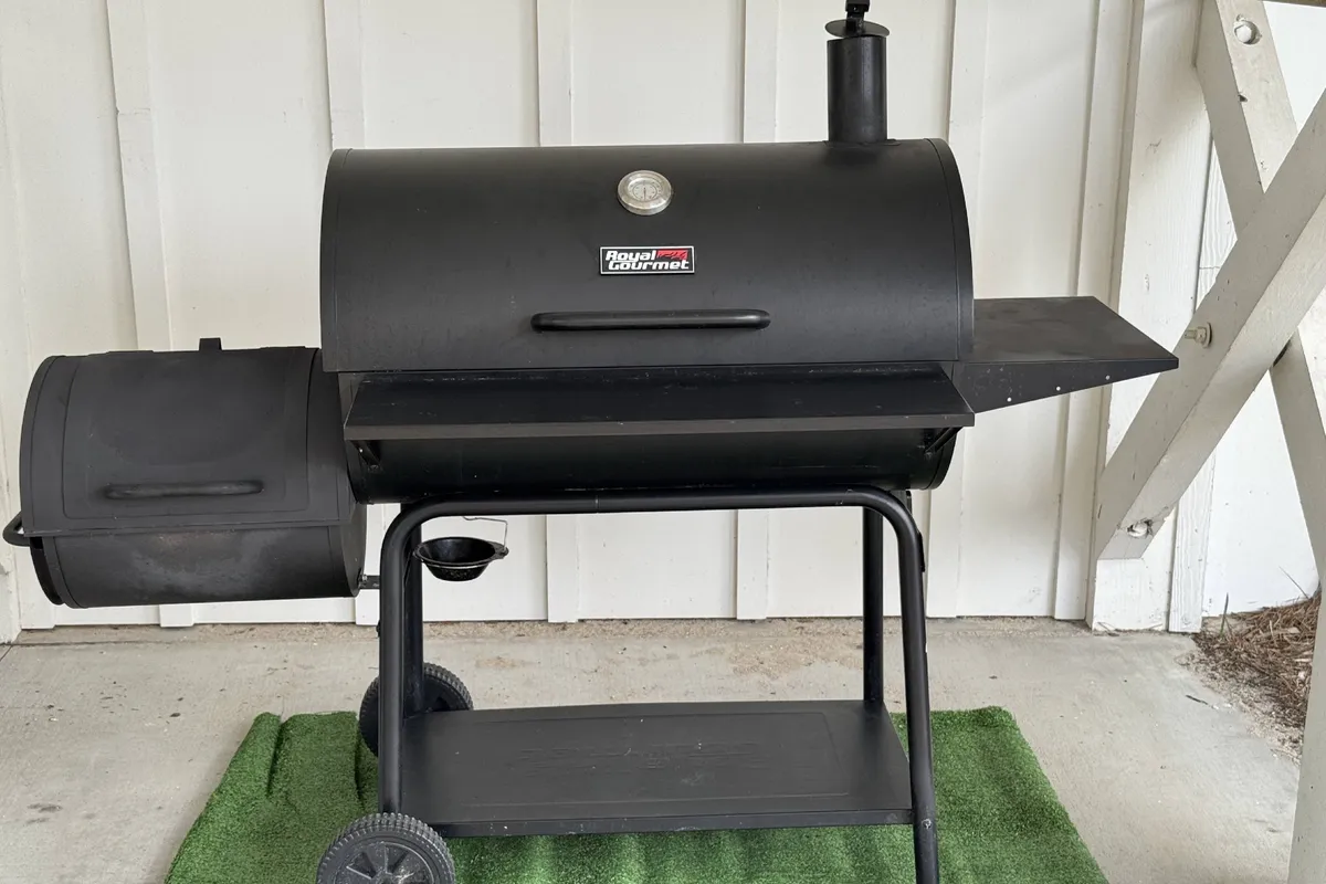 This image centers on a smoker grill and grill accessories set up on a green mat. The exterior scene hints at a casual, social atmosphere perfect for outdoor cooking and shared meals.