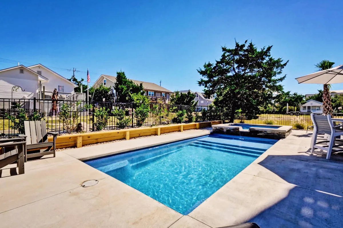 This exterior image features a sparkling swimming pool surrounded by inviting patio furniture, a partially shaded seating area under a tree, and a fenced yard for privacy. The relaxed mood is heightened by ample space for sunbathing and gathering with friends or family, evoking a resort-like atmosphere.