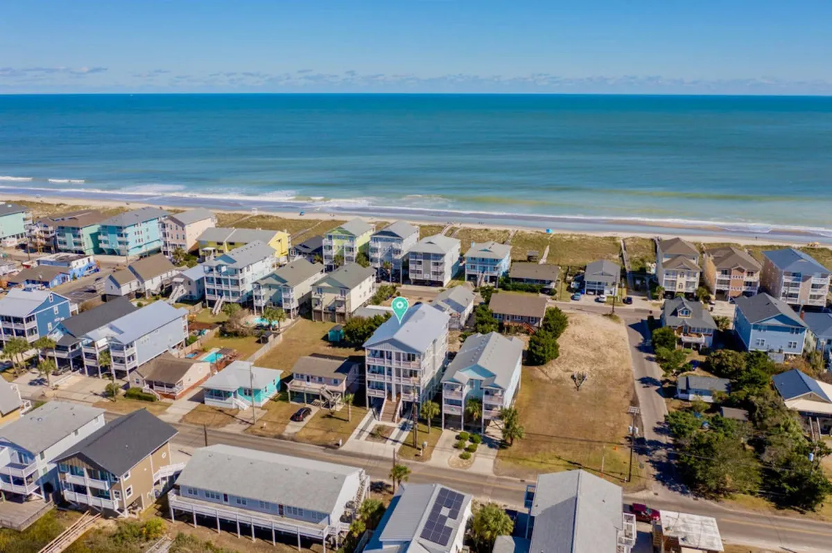 A sweeping exterior view showcasing oceanfront homes directly facing the beach, with the neighborhood layout visible and the sandy shoreline right in front. The image exudes a relaxed atmosphere, suggesting easy access to beach activities.