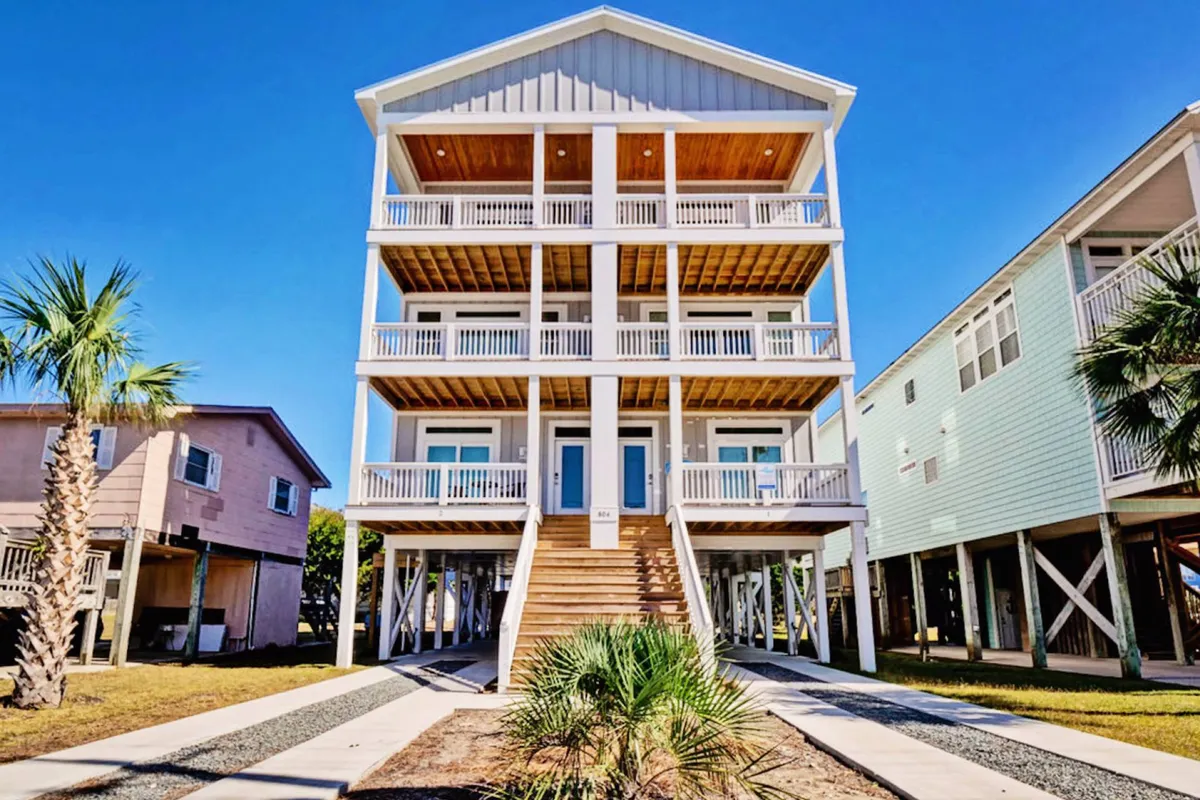 A cheerful exterior shot of Blue Oasis showing its three-story structure, inviting balconies with railings, wooden steps, and palm trees. The scene suggests accessibility to the beach and offers a glimpse of the home's outdoor lifestyle, including the spacious decks mentioned in the section.