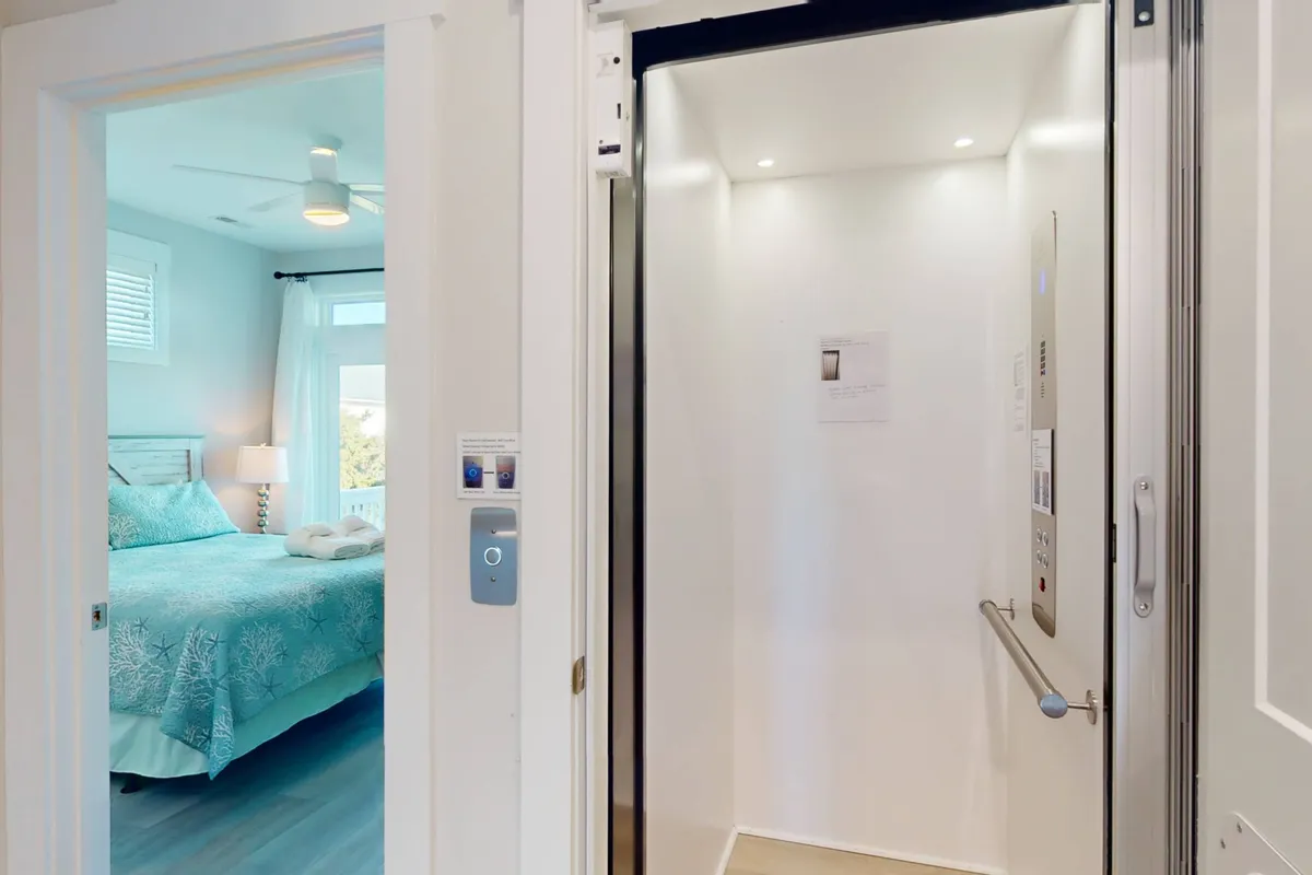 An interior hallway with a visible elevator next to a doorway to a cozy bedroom, featuring a neatly made bed with decorative pillows. The elevator doors are open, clearly showcasing this essential amenity for guests who prefer to skip the stairs.