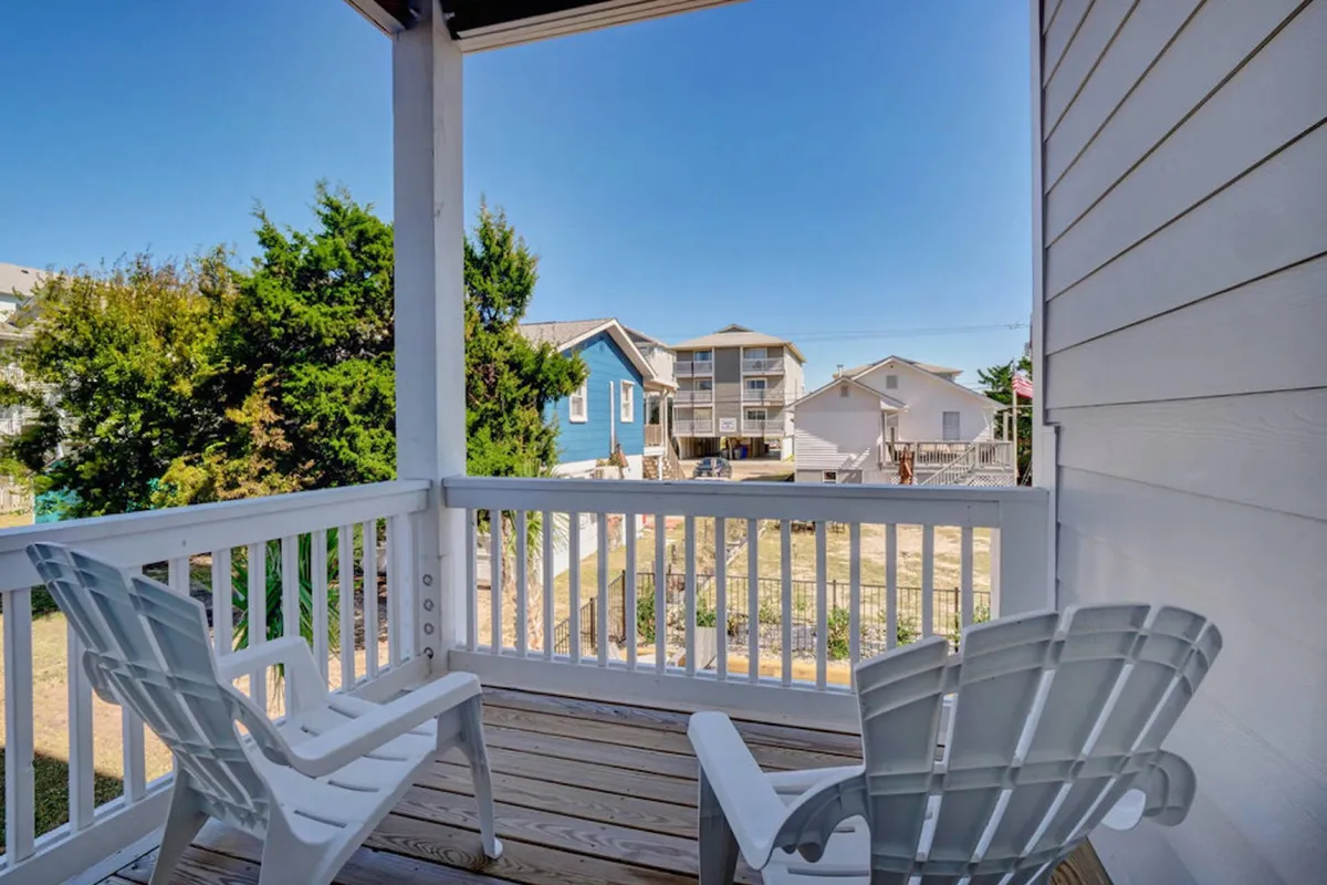 A tranquil outdoor sitting area with white Adirondack chairs set amidst green foliage, colorful beach houses in the background, and a welcoming, open-air feel. The scene conveys relaxation and a neighborhood atmosphere.
