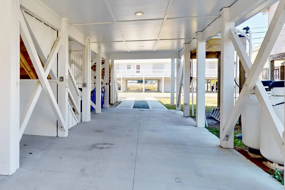 A ground-level view of the spacious parking area under the house, showing multiple garage doors and support beams, with an open pathway that provides direct, easy access to the main entrance. The area appears functional and welcoming, perfect for parking multiple vehicles and storing outdoor gear.